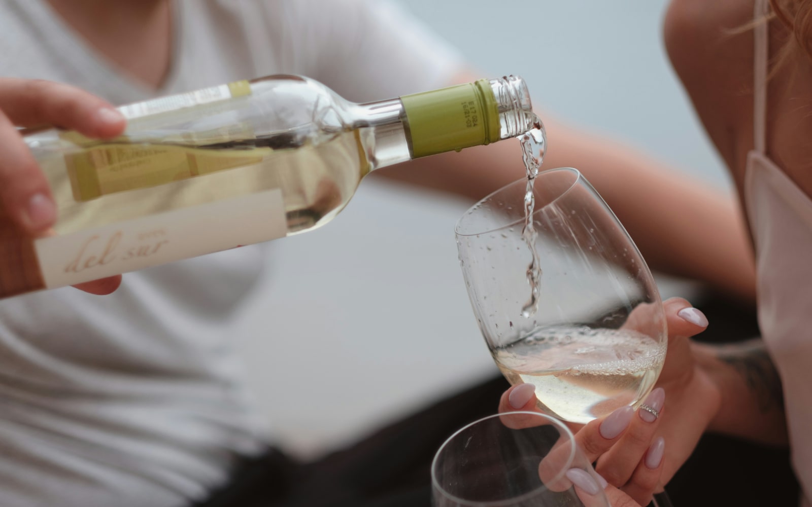 Close-up of a person pouring white wine from a bottle into a glass held by another person, highlighting the wine's flow and the intimate setting.