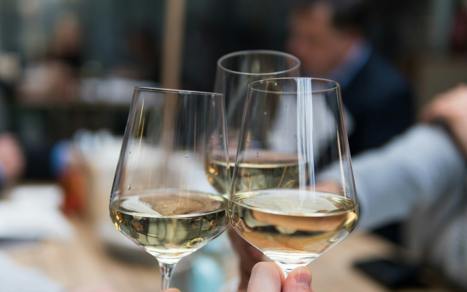 Close-up of three people clinking glasses of white wine in a celebratory toast, with a blurred background of a social setting.