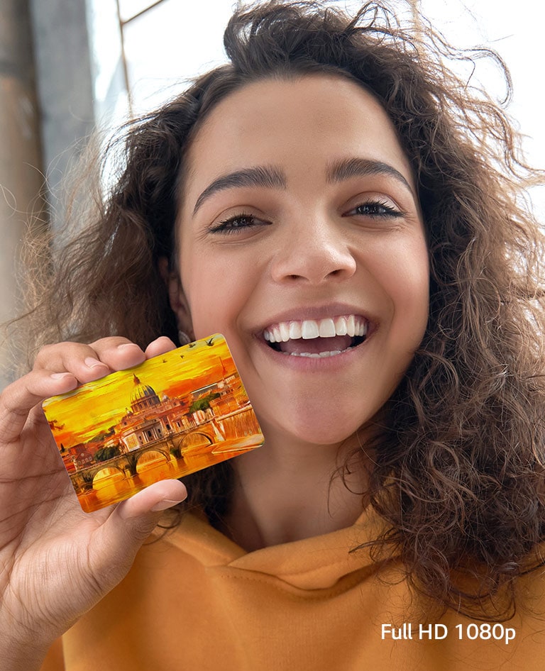 A woman is holding a yellow card in her right hand, facing forward and smiling brightly.