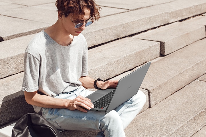 L'immagine mostra un giovane uomo seduto su gradini di cemento all'aperto, che utilizza un computer portatile grigio. L'uomo sembra concentrato sul suo lavoro o sulla navigazione, evidenziando la portabilità del computer portatile e la sua idoneità all'uso in movimento.