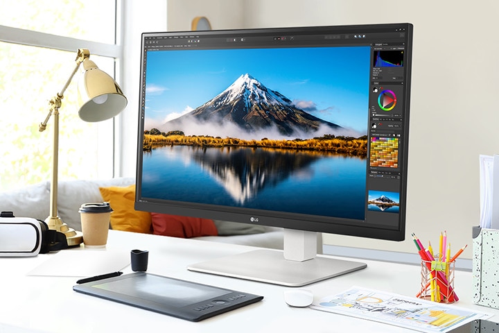 On the desk, there is an ultrafine monitor displaying Photoshop work. Next to the monitor, there is a desk lamp, a mouse, paper, and colored pencils.