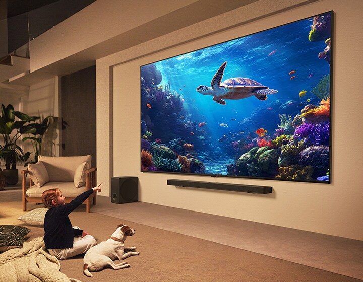 Een gigantische LG QNED TV en LG Soundbar zijn op de wand gemonteerd. Een klein meisje en haar puppy kijken naar een onderwaterbeeld op de TV. Het grootste formaat van de TV dompelt de kijker nog meer onder in de content.