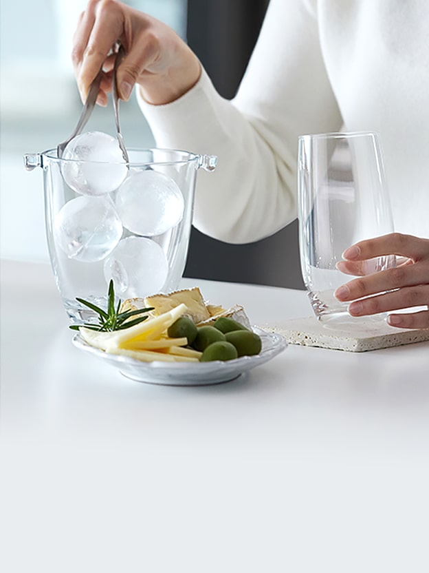 Een Craft Ice Maker die ronde ijsballen maakt, terwijl iemand met een tang een ijsbal in een glas legt naast een bord met snacks.