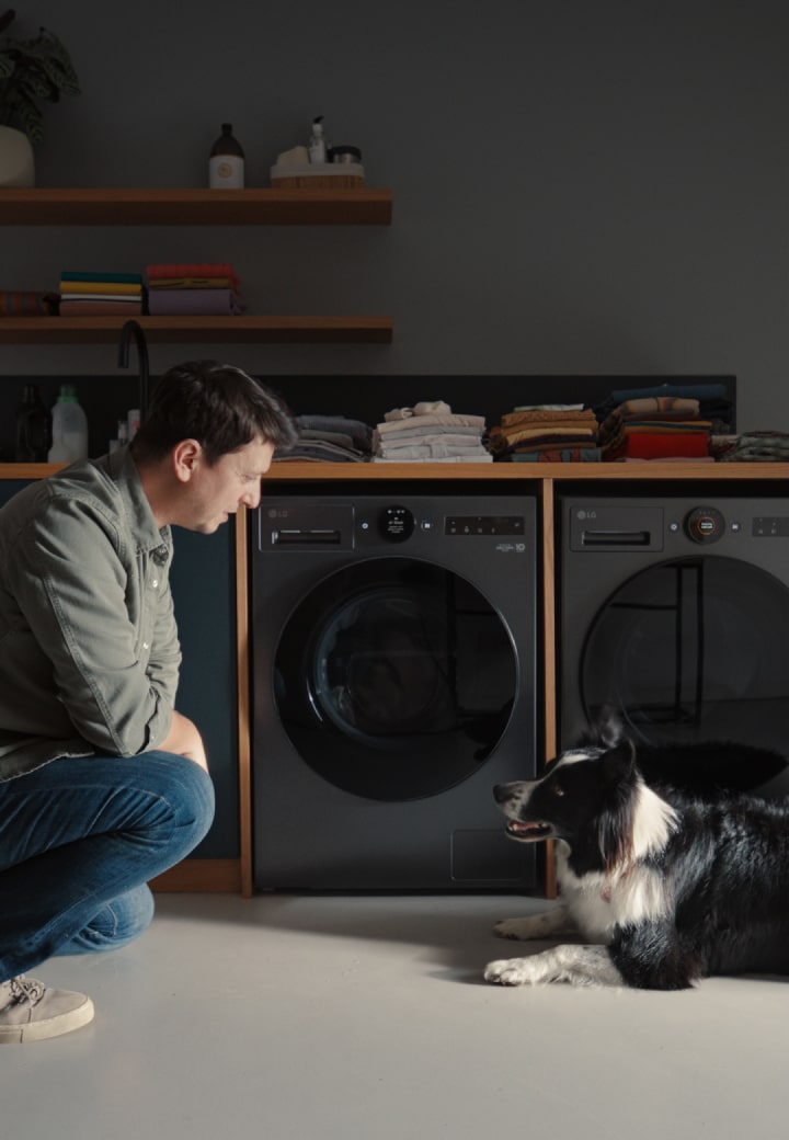 Een man in een olijfgroen shirt en jeans knielt en kijkt naar een zwart-witte hond voor een zwarte LG wasmachine.