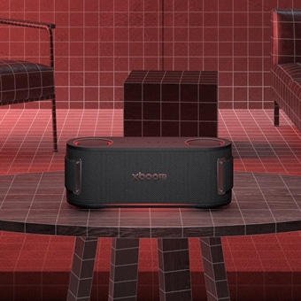 LG XBOOM-speaker geplaatst op een tafel in een rood getinte kamer met wanden in rasterpatroon en moderne meubels.