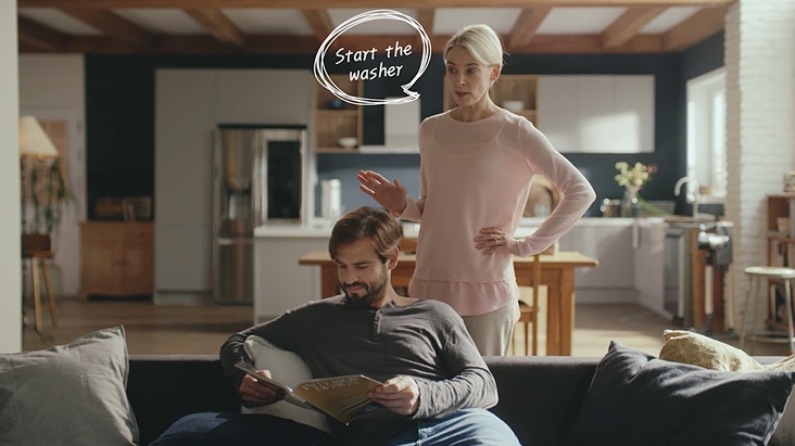 In een moderne slimme keuken gebruikt een vrouw een spraakopdracht om de wasmachine te starten met LG ThinQ AI, terwijl een man op de bank op de achtergrond zit te lezen.