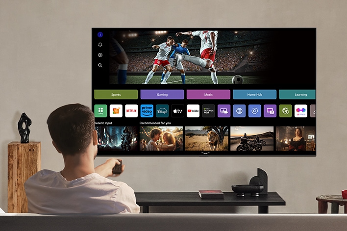 Een persoon op de bank die een afstandsbediening vasthoudt. Een LG AI TV aan de muur met LG webOS op het scherm.