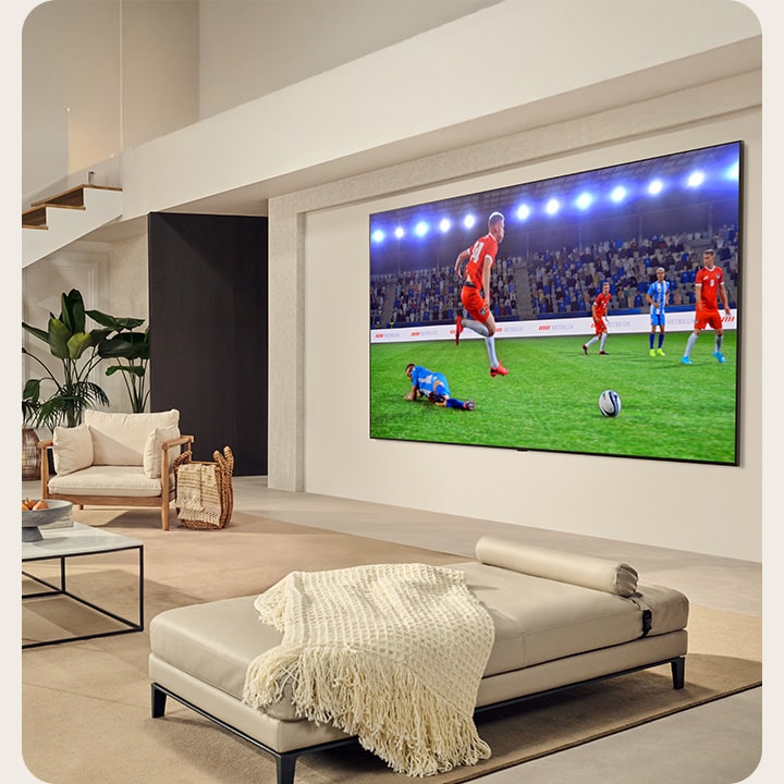 Een familie zit in hun woonkamer. Ze hebben een op de wand gemonteerde LG TV met een ultragroot scherm dat bijna net zo groot is als de hele wand. Ze kijken naar een spannende voetbalwedstrijd op het grote en meeslepende scherm.