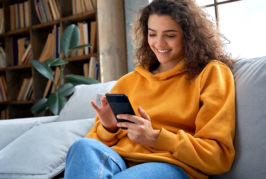 Een vrouw in een gele hoodie glimlacht terwijl ze op de bank naar haar smartphone kijkt.