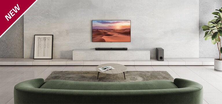 De tv is opgehangen aan de witte pilaar in het midden van de woonkamer. Hieronder staat de LG Soundbar op de witte plank. Op de vloer rechts onder de soundbar staat een draadloze subwoofer. Achter de groene bank zijn links en rechts op de salontafel twee achterspeakers geplaatst. In de linkerbovenhoek staat NEW mark.