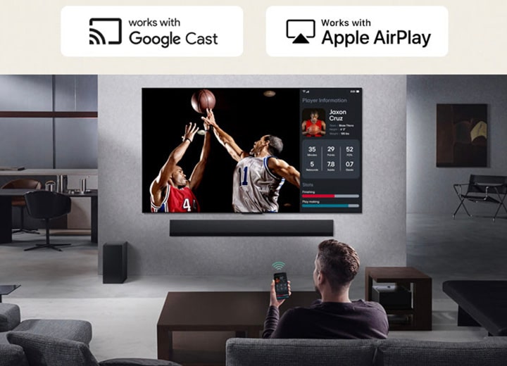 Uma pessoa na sala de estar segurando o telefone. No telefone há um ícone de transmissão mostrando que a tela do telefone está sendo espelhada na TV. Na TV há um jogo de basquete e ao lado há uma tela espelhada, mostrando as estatísticas dos jogadores.