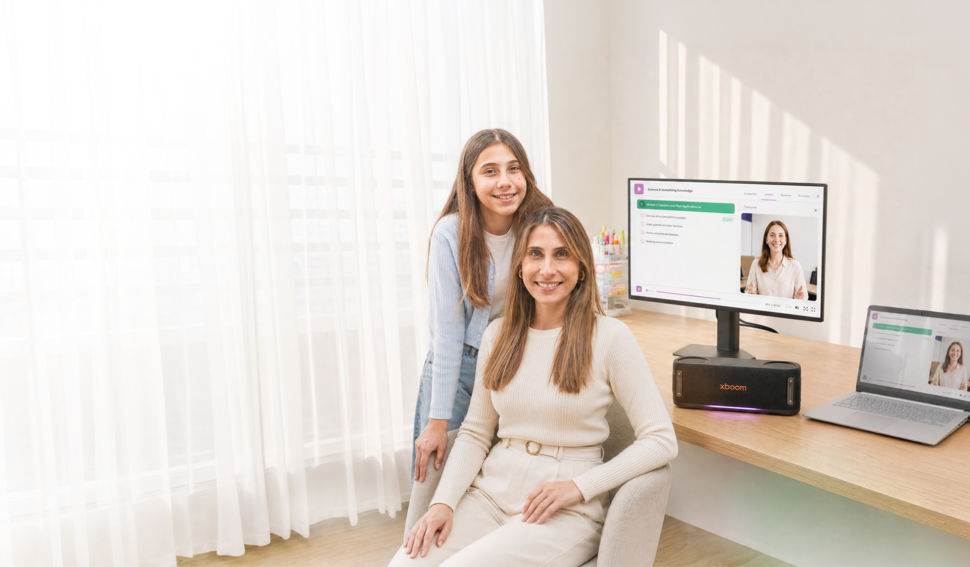Mãe sentada e filha em pé sorrindo em uma mesa iluminada com monitor LG, caixa de som xboom e notebook