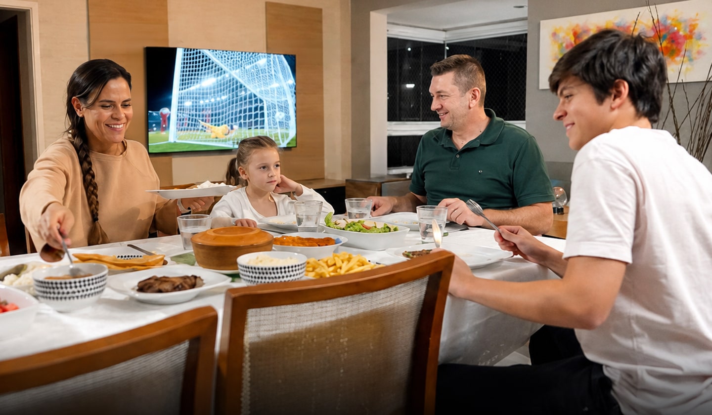 Família à mesa com um jogo de futebol passando na TV ao fundo