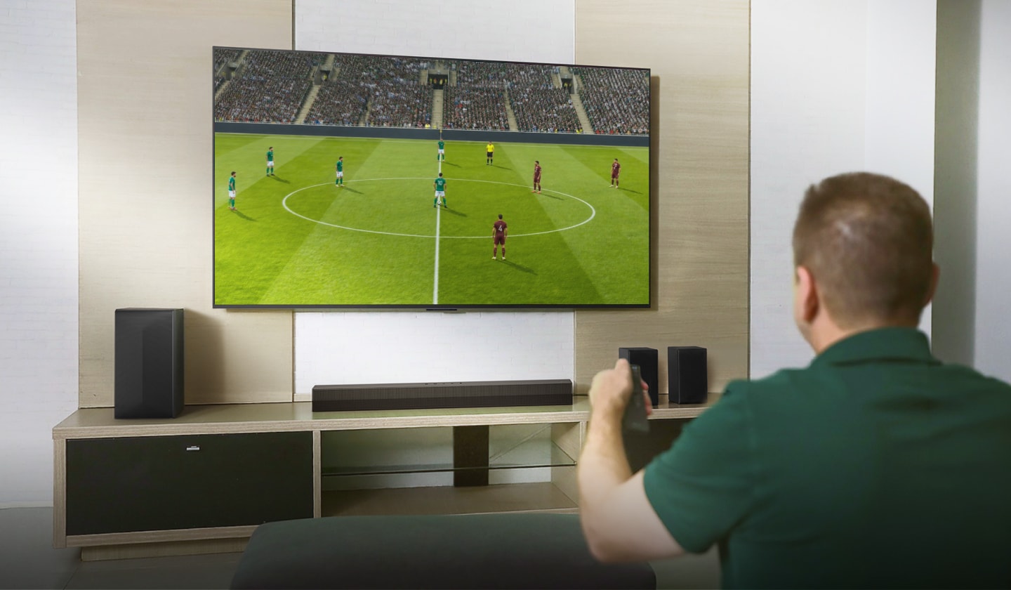 Marcos segura o controle remoto enquanto assiste a um lance de futebol em uma TV grande na sala de estar