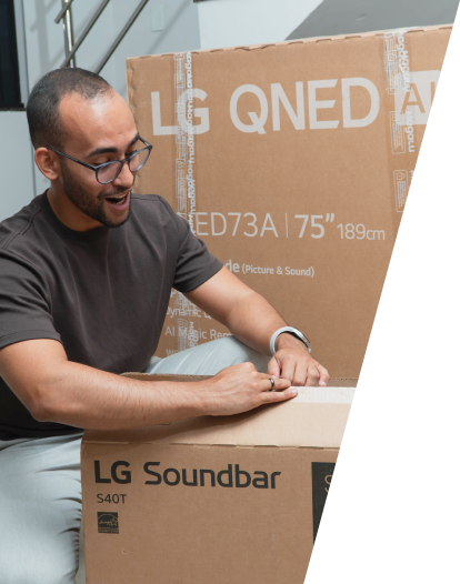 Rafinha animado ao abrir a caixa de uma nova Soundbar LG com a caixa de uma LG QNED TV ao fundo