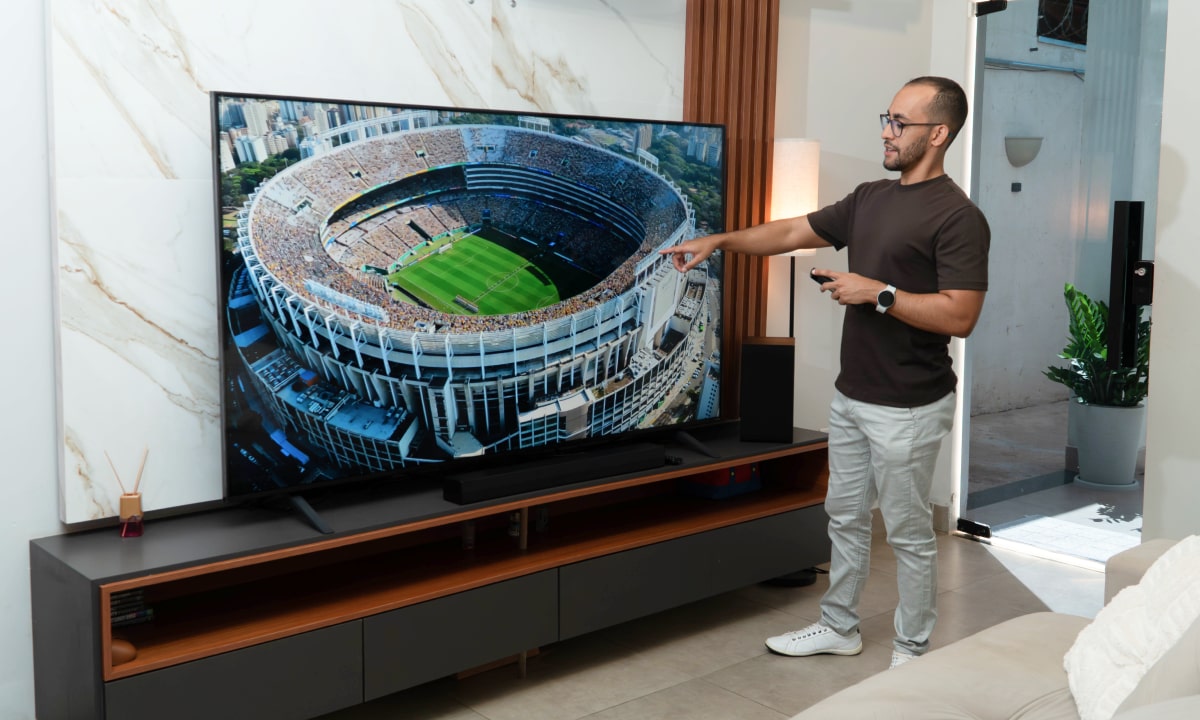Rafinha em pé e apontando para uma TV grande mostrando um estádio de futebol lotado, enquanto segura um controle remoto