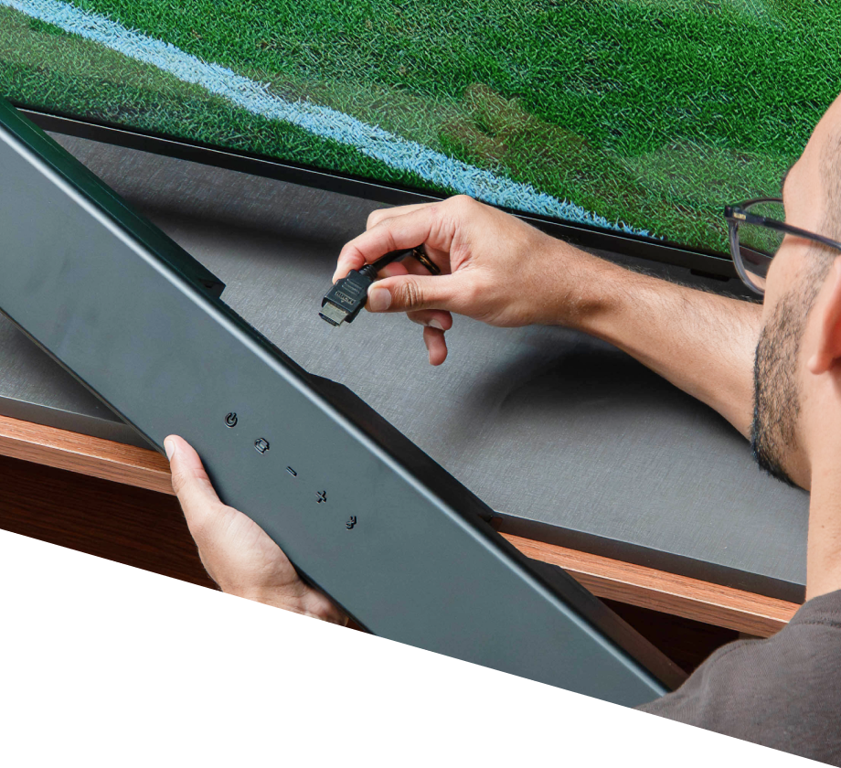 Close-up do Rafinha conectando um cabo HDMI a uma soundbar em frente a uma tela de TV