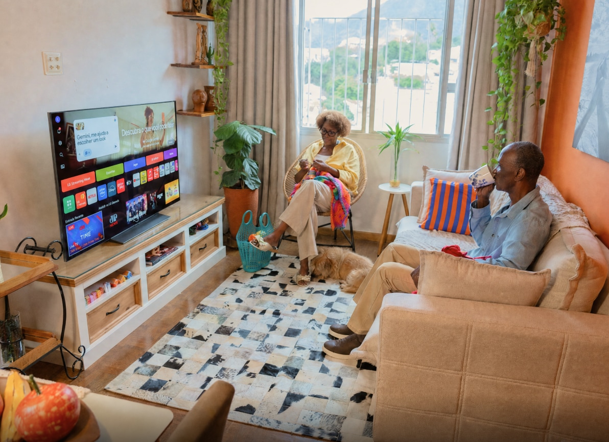 Sandra faz crochê na poltrona enquanto Manoel segura uma caneca no sofá, com a tela inicial da TV LG OLED ao lado.