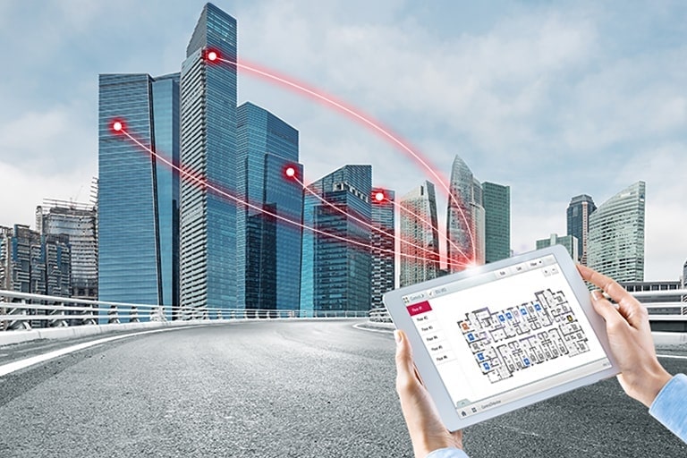 Uma pessoa segura um tablet exibindo uma solução de controle. Quatro linhas vermelhas se estendem do tablet, conectando-se aos edifícios à esquerda.