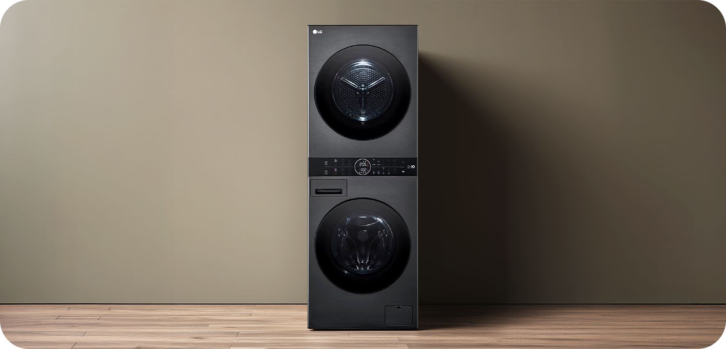 A black washer-dryer tower unit is centered against a beige wall and wooden floor background. The dryer is positioned on top and the washer on the bottom, with a circular control dial panel in the middle.