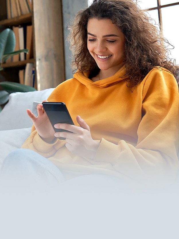 Uma mulher de moletom amarelo sorri enquanto olha para o smartphone no sofá.