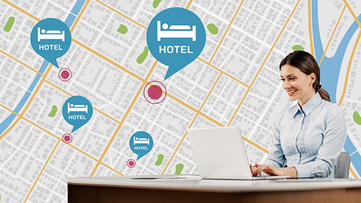 Uma mulher sentada à mesa está usando seu PC para gerenciar as TVs do hotel em diferentes locais.	