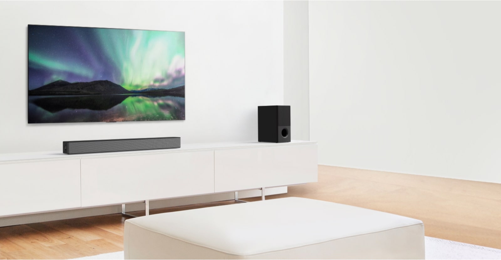 TV e Sound bar numa sala de estar branca com um sofá branco ao centro. Os alto-falantes estão em ambas as extremidades do sofá.