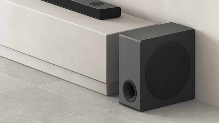 O subwoofer está colocado ao lado de um rack bege na sala de estar.