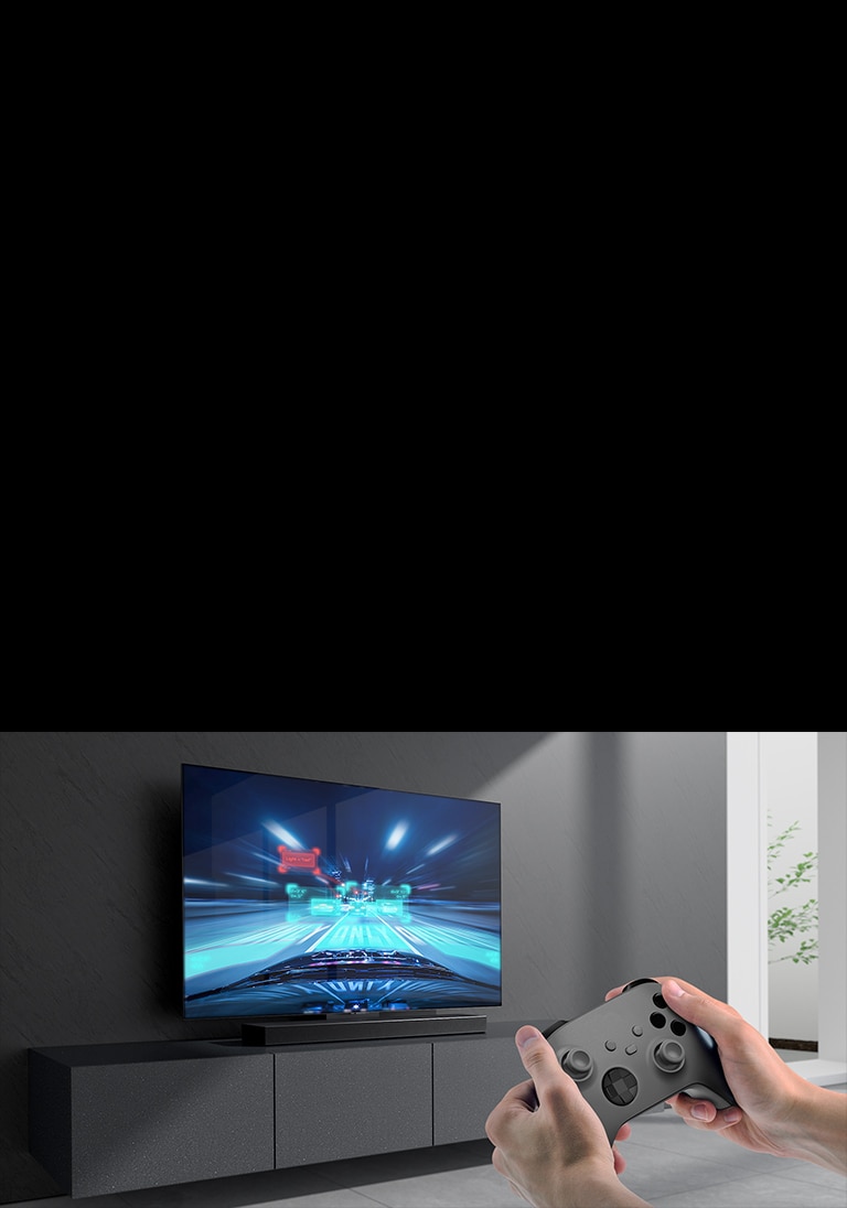A barra de som está colocada sobre um rack, e a cena de um jogo de corrida é mostrada na TV conectada a ela. Um console de jogo está no canto inferior direito da imagem, segurado por duas mãos.