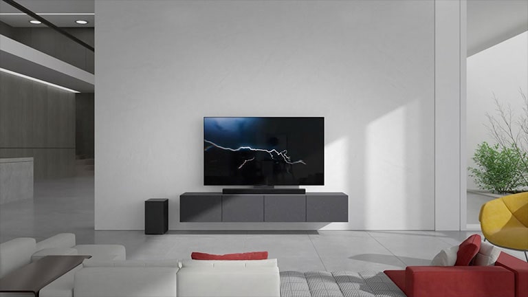 A barra de som e a TV estão sobre um rack cinza na sala de estar. Um subwoofer preto sem fio está no chão, do lado esquerdo, e há luz solar entrando pelo lado direito da imagem. Um longo sofá branco e vermelho está de frente para a TV e a barra de som.