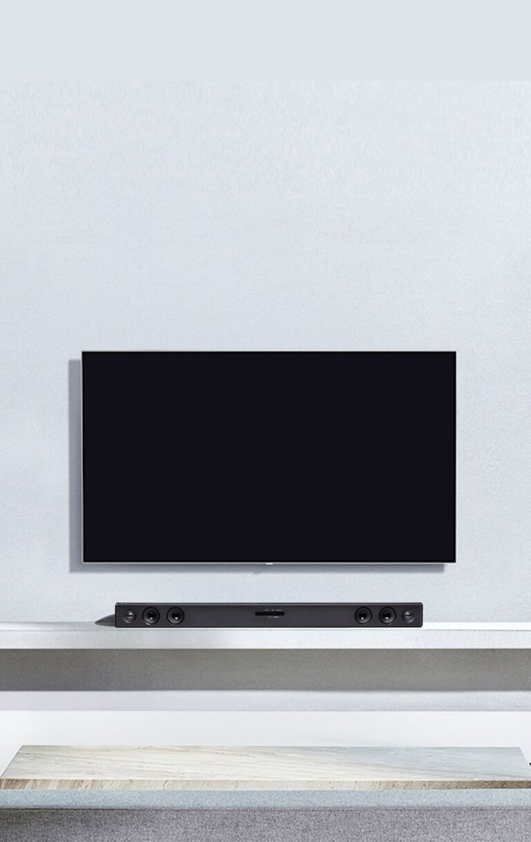 A TV LG está pendurada numa parede cinza. E a Sound Bar LG SQC2 está colocada na prateleira da sala de estar. O subwoofer está colocado ao lado da prateleira. A imagem ilustra que a sound bar combina com a TV LG e complementa a decoração.