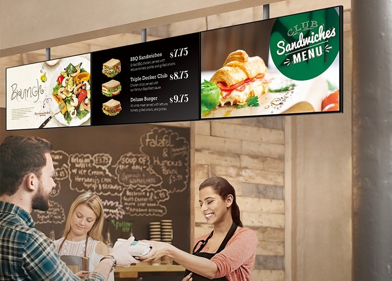 Colaboradora de uma lanchonete entrega sanduíche ao cliente. O monitor SM5J está instalado acima deles, exibindo opções de sanduíches em promoção.
