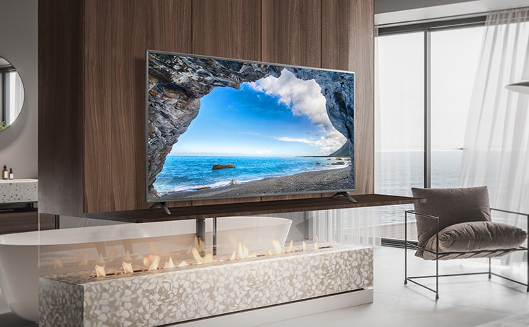 Em um quarto simples com vista para o mar, há uma TV em uma prateleira de parede. A paisagem azul do mar aparece brilhante e nítida na tela da TV.