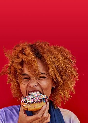  Pessoa com cabelo cacheado segurando um donut rosa com granulados coloridos em frente a um fundo vermelho.
