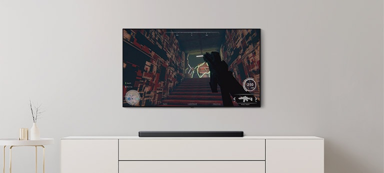Há uma TV e uma barra de som na sala de estar. Um jogo FPS aparece na tela da TV e o canal da TV muda para um jogo de futebol. (reproduza o vídeo)