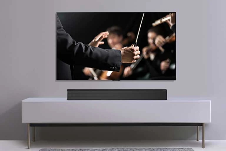 Uma TV é exibida em uma parede cinza e o LG Soundbar abaixo dela em uma prateleira cinza. A TV mostra um maestro conduzindo uma orquestra.