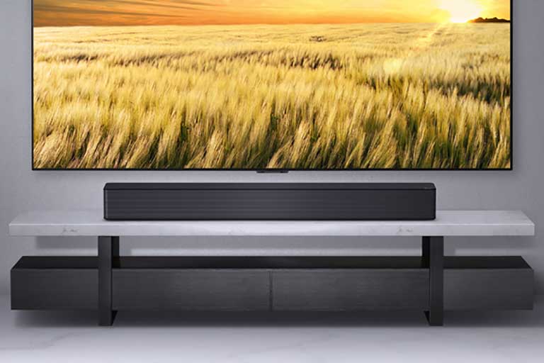 Uma TV é exibida em uma parede cinza e o LG Soundbar abaixo dela em uma prateleira cinza. Disco Blue-Ray abaixo da prateleira.