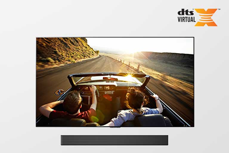 TV e Sound bar juntos em vista completa. A TV mostra um casal em um carro conversível numa estrada, rumo ao pôr do sol.