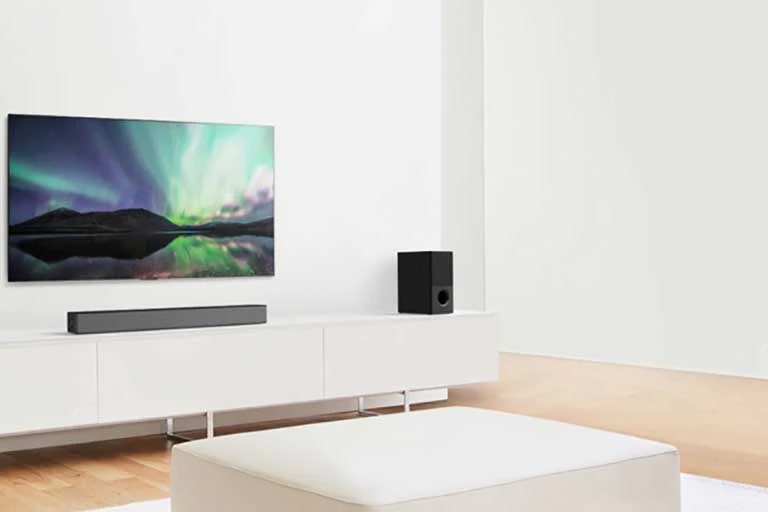 TV e Sound bar numa sala de estar branca com um sofá branco ao centro. Os alto-falantes estão em ambas as extremidades do sofá.