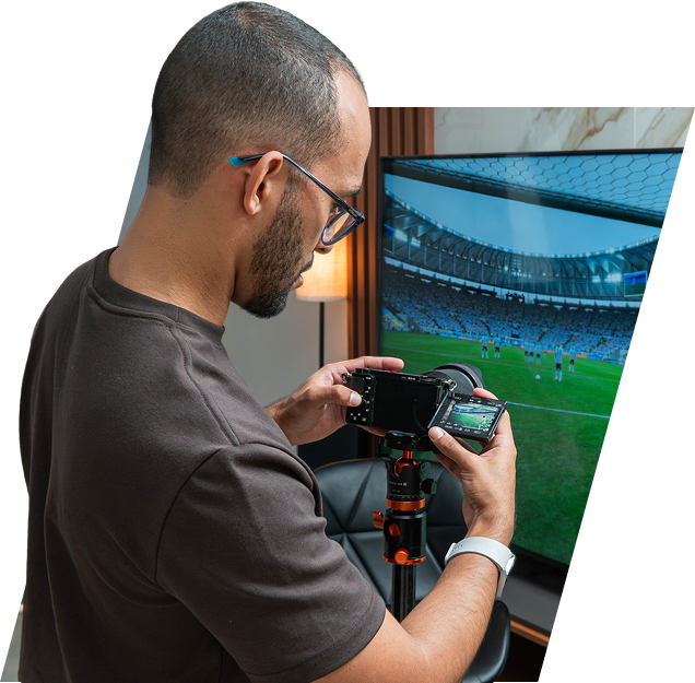 Rafinha usando uma câmera em um tripé para fotografar um jogo de futebol exibido na tela da TV