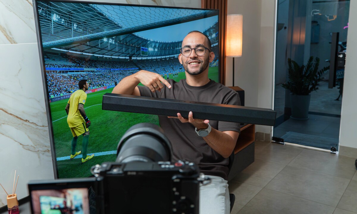 Rafinha sorrindo e apontando para uma soundbar que ele está segurando, com uma TV mostrando um jogo de futebol ao fundo