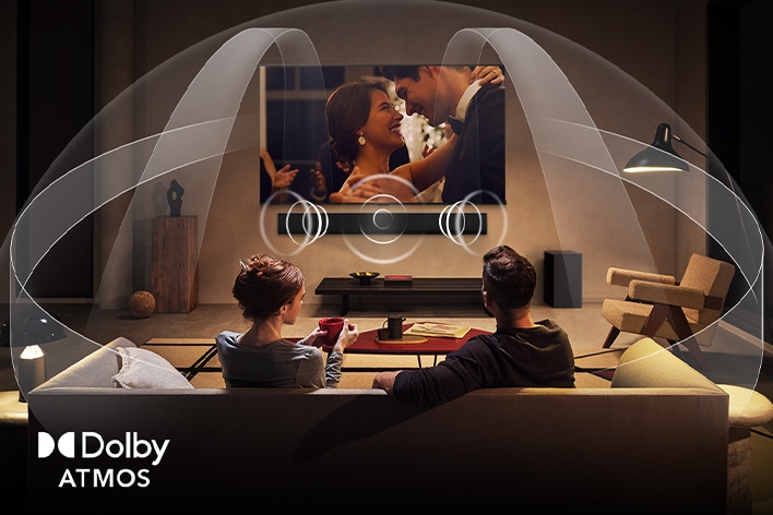 Dimly lit, cozy living room with a couple watching a movie on their LG TV with LG Soundbar. Graphics show how Dolby Atmos fills the whole space with surround sound from all directions. Dolby Atmos logo is at the bottom corner.
