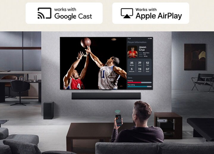 Person in their living room holding their phone. On the phone is a transmitting icon showing that the phone screen is being mirrored on the TV. On the TV is a basketball game to the side is the mirrored screen, showing player statistics.