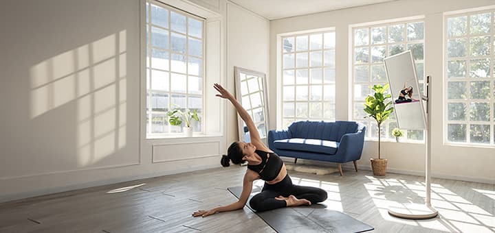 Woman in her spacious home doing yoga. Beside her is the LG StanbyME TV on a stand. It's in the vertical orientation and is playing the yoga instructional video that the woman is following.