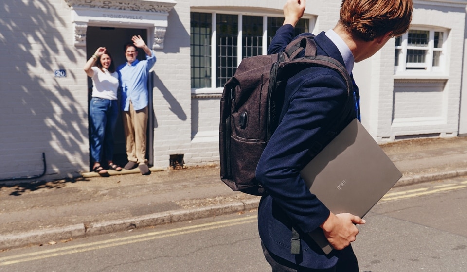 Student carrying a laptop and backpack while waving goodbye to his parents before school.