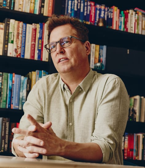 A man wearing glasses sits indoors in front of a bookshelf, gesturing with his hands as if explaining something.