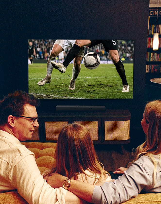 A man wearing glasses laughs warmly in a dimly lit living room, a glowing lamp visible beside him.