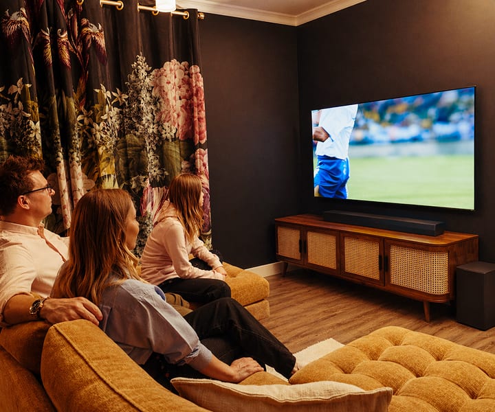 A family of three sits together on a sofa in a cozy, dimly lit living room, watching a soccer match on a large LG OLED TV. The room features dark walls, floral curtains, a wooden media console, and speakers, creating a warm and intimate atmosphere.