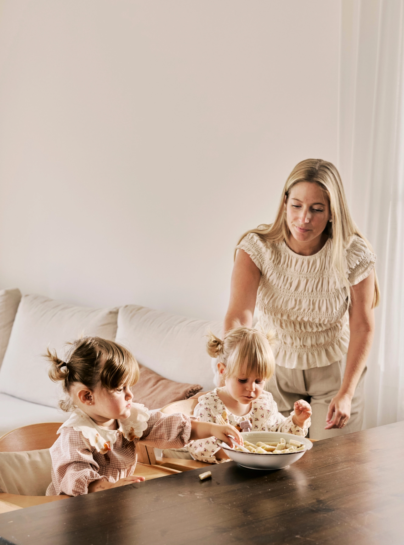 A mother watching over her toddler twins as they eat snacks at a wooden dining table.
