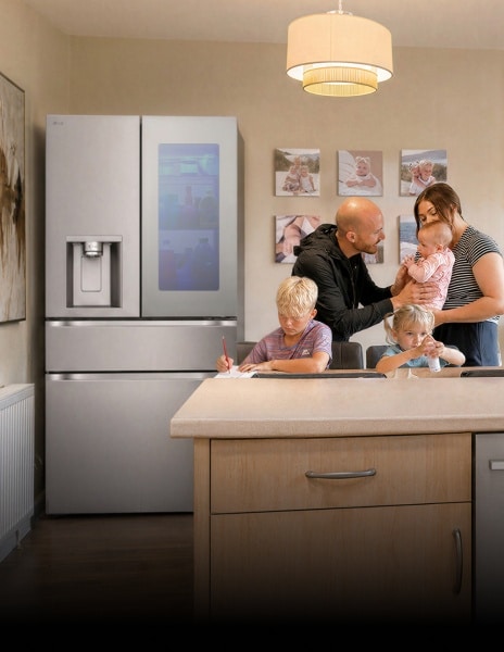 A family gathers in a kitchen near a stainless steel LG refrigerator—a boy writes at the dining table, a young girl sits beside him with a small bottle, and parents stand together holding a baby.